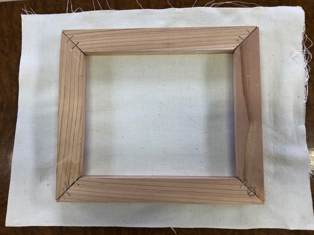 木枠を布の上に置き、周囲に約5cmの余白（掴みしろ）を持たせて布を切り出している様子。木枠よりも一回り大きく布をカットする工程の解説。