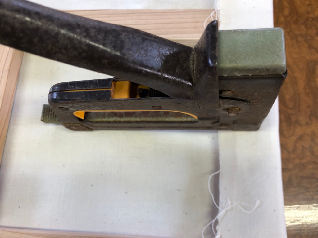 アイロンをかけた布の中央に木枠を配置し、最初の一箇所をタッカーで固定している様子。布張り作業の開始地点を示す画像。