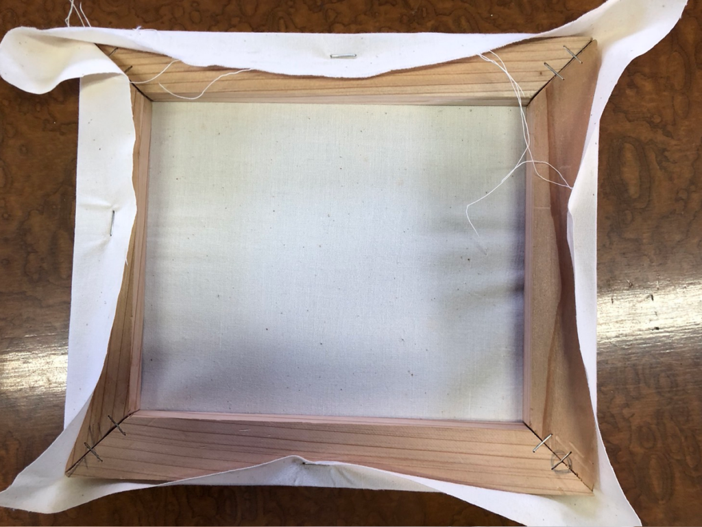 長辺に続いて短辺の中央もタッカーで固定し、布に十字型の張りがかかっている様子。対角線上に引っ張って均一なテンションを作る工程。